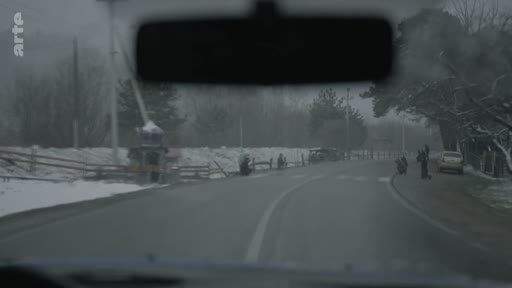 Vue depuis l'intérieur d'une voiture circulant sur une route bordée de neige dans un village rural, avec des piétons marchant sur le bas-côté.