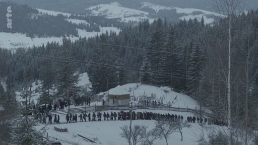 Une longue file de personnes vêtues de vêtements sombres est rassemblée dans un cimetière enneigé en montagne, lors d'une cérémonie funéraire. La scène se déroule dans un paysage hivernal boisé, typique des régions montagneuses.
