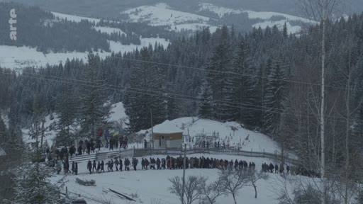 Une longue file de personnes se recueille lors d'une cérémonie d'enterrement dans un cimetière situé en zone montagneuse enneigée. La scène montre une foule rassemblée pour un dernier hommage dans un paysage hivernal.
