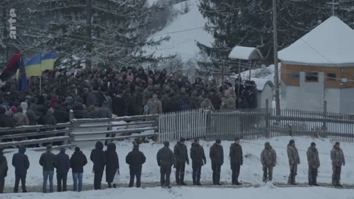 Une foule de personnes, dont certaines en tenue militaire, assiste à une cérémonie funéraire en extérieur dans un paysage enneigé, avec des drapeaux ukrainiens visibles. La scène se déroule près d'une église en bois dans une zone rurale, illustrant un hommage solennel.