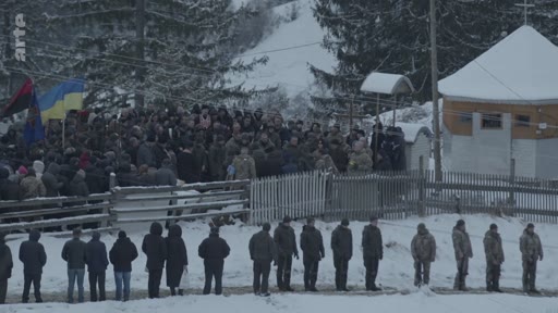 Une foule de personnes, dont des hommes en uniforme militaire, est rassemblée dans un paysage enneigé en Ukraine pour une cérémonie funéraire. Des drapeaux ukrainiens sont visibles près d'une petite église en bois, illustrant un hommage solennel.