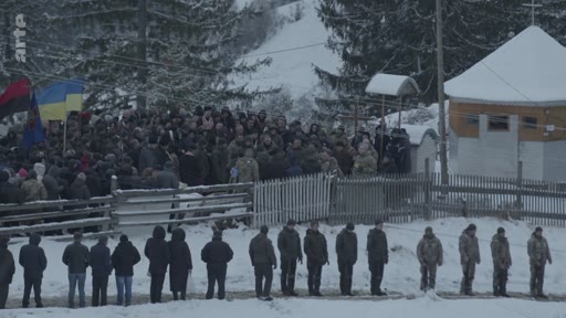 Une foule de personnes, dont des militaires en uniforme, est rassemblée pour une cérémonie funéraire en extérieur dans un paysage enneigé en Ukraine. On distingue des drapeaux ukrainiens et des drapeaux rouge et noir, symboles de la résistance ukrainienne, lors de cet hommage solennel.