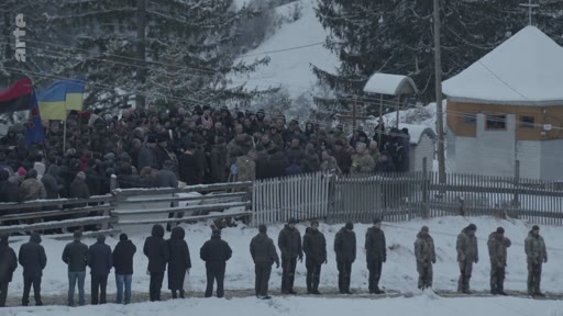 Une foule de personnes assiste à une cérémonie funéraire en extérieur dans un paysage enneigé en Ukraine, avec des drapeaux ukrainiens visibles. Des hommes sont alignés au premier plan lors de cet hommage solennel.