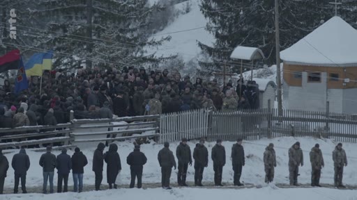 Une foule nombreuse et des soldats en uniforme assistent à une cérémonie funéraire en plein air dans un paysage enneigé, avec des drapeaux ukrainiens visibles. La scène se déroule près d'une petite église en bois, marquant un moment de recueillement collectif.