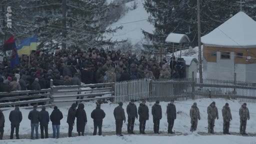 Une foule assiste à une cérémonie funéraire en extérieur dans un village ukrainien enneigé, avec des drapeaux ukrainiens et des membres de la communauté réunis près d'une église. Des hommes en uniforme et en civil sont alignés dans la neige pour rendre hommage lors de cet enterrement.