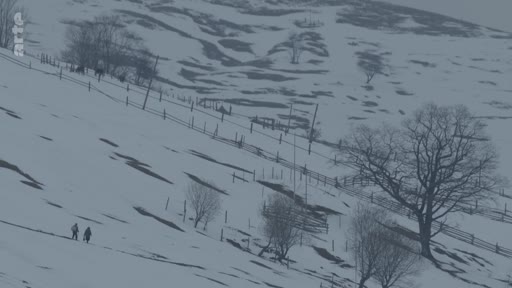 Deux personnes traversent un paysage montagneux enneigé, marqué par des clôtures et des arbres dénudés, illustrant la vie quotidienne dans une région rurale en hiver.