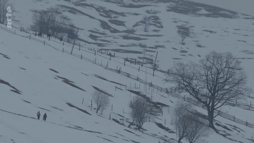 Deux personnes traversent un paysage montagneux enneigé et désolé, illustrant la vie quotidienne dans une zone rurale isolée ou en contexte de conflit.