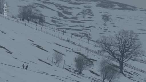 Deux individus traversent un terrain montagneux recouvert de neige, marqué par des clôtures en bois et des arbres dénudés, illustrant la vie quotidienne dans une région rurale isolée.