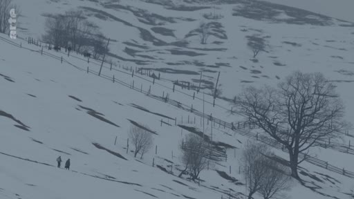 Deux personnes traversent un paysage montagneux enneigé et désolé, marqué par des clôtures en bois, illustrant la vie quotidienne dans une zone rurale isolée.