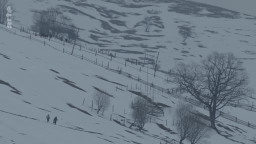 Deux personnes traversent un paysage montagneux enneigé et isolé, marqué par des clôtures en bois et des arbres dénudés, illustrant la vie locale dans une région rurale en hiver.