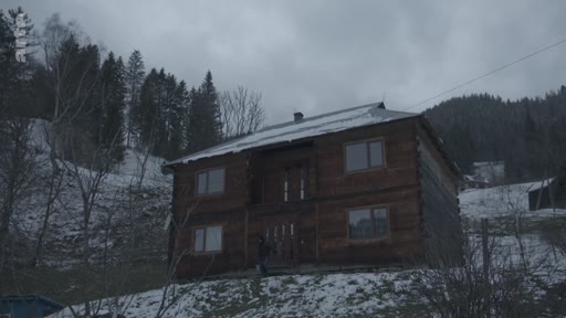 Vue extérieure d'une maison en bois traditionnelle située dans un village de montagne enneigé, illustrant les conditions de vie difficiles en hiver.
