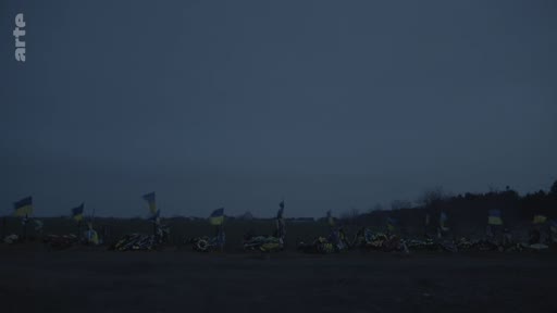 Vue d'ensemble d'un site commémoratif marqué par de nombreux drapeaux ukrainiens plantés dans le sol, illustrant les conséquences de la guerre en Ukraine. L'image, prise à la tombée de la nuit, souligne une atmosphère de recueillement et de deuil.