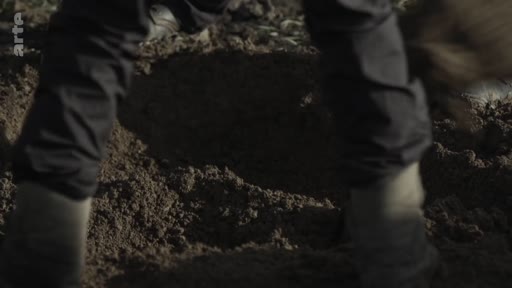 Gros plan sur les jambes et les bottes d'une personne en train de creuser le sol dans le cadre d'une opération d'exhumation et de recherche de restes humains.