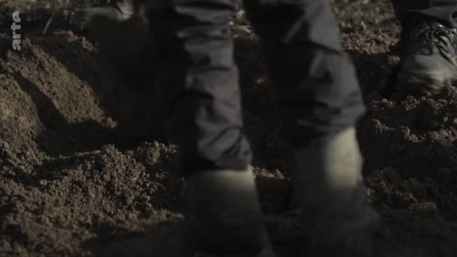 Gros plan sur les jambes et les chaussures de personnes marchant sur un terrain terreux lors d'une opération d'exhumation ou de recherche médico-légale.