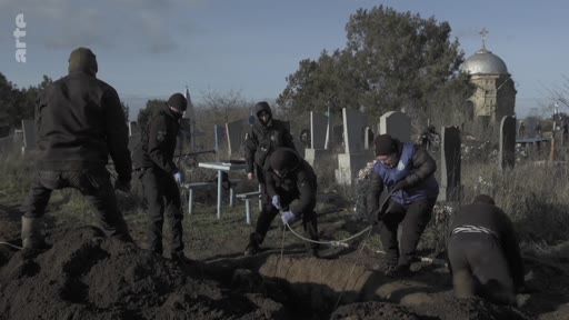 Des individus en vêtements sombres et gants de protection enterrent un corps dans un cimetière, avec une église à coupole en arrière-plan. Cette scène illustre les opérations d'inhumation dans un contexte de conflit ou de crise humanitaire.