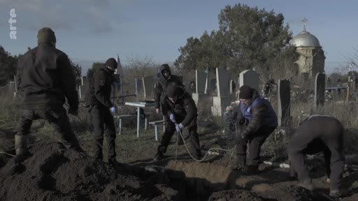 Des hommes en vêtements sombres et gants de protection enterrent un cercueil dans un cimetière en Ukraine, près d'une église orthodoxe. Cette scène illustre les conséquences tragiques du conflit en Ukraine et le travail des équipes chargées des inhumations.