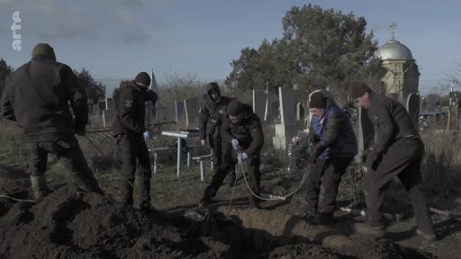 Des hommes en vêtements sombres travaillent ensemble pour enterrer un corps dans un cimetière, avec une église en arrière-plan. Cette scène illustre les opérations d'inhumation dans un contexte de conflit ou de crise humanitaire.