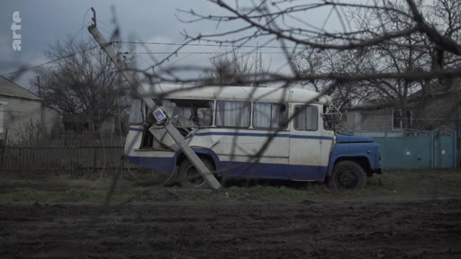 Un vieux bus scolaire ou de transport public, partiellement détruit et renversé, gît dans un champ boueux près d'habitations rurales, illustrant les conséquences de la guerre sur les infrastructures civiles.