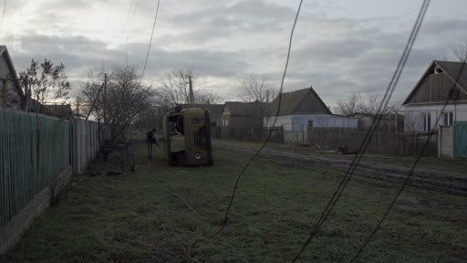 Un soldat en uniforme patrouille dans une rue résidentielle d'un village ukrainien, passant devant la carcasse calcinée d'un véhicule militaire. Cette scène illustre les conséquences des combats dans les zones rurales lors du conflit en Ukraine.