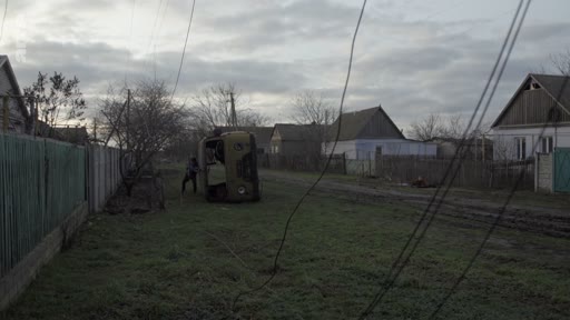 Un soldat en uniforme militaire examine les restes d'un véhicule blindé calciné dans une rue résidentielle d'un village en Ukraine, illustrant les conséquences des combats sur le terrain.