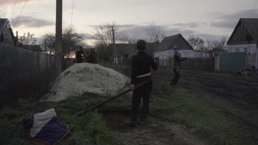 Deux individus, dont un homme portant un casque, effectuent des travaux manuels dans un jardin à proximité d'habitations rurales en Ukraine, dans un contexte de conflit armé.