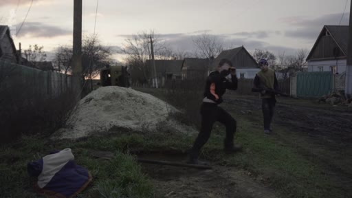 Deux soldats en tenue de combat patrouillent dans une rue d'un village ukrainien lors d'une mission de surveillance au crépuscule. L'un des soldats utilise des jumelles pour observer les environs près d'un tas de sable.