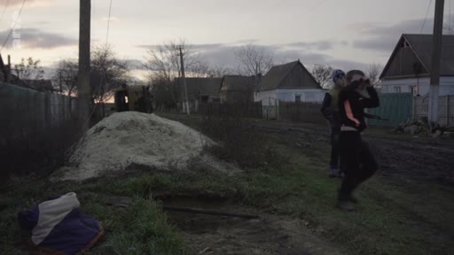 Des soldats ukrainiens en patrouille dans une zone rurale, observant les environs avec des jumelles près d'un monticule de terre. La scène illustre la présence militaire dans les villages ukrainiens pendant le conflit en Ukraine.