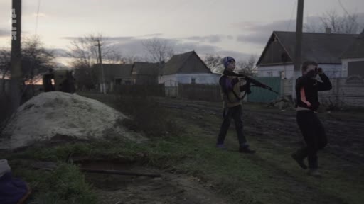 Deux jeunes hommes, dont l'un porte une arme, patrouillent dans une rue d'un village ukrainien lors du conflit en Ukraine. La scène illustre la présence de civils ou de volontaires armés dans les zones rurales touchées par la guerre.