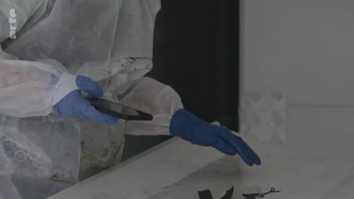 Un expert en combinaison de protection et gants en latex examine des objets personnels sur une table lors d'une enquête médico-légale. La scène illustre le travail de police scientifique ou d'analyse de preuves sur une scène de crime.