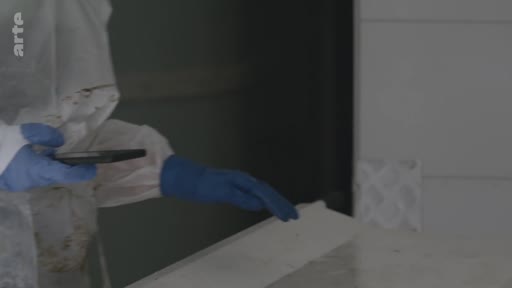 Un enquêteur portant une combinaison de protection intégrale et des gants bleus examine une surface plane à l'aide d'un téléphone portable, dans le cadre d'une investigation médico-légale.