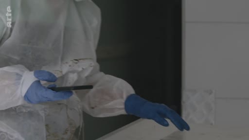 Une personne portant une combinaison de protection intégrale et des gants en latex bleu examine un téléphone portable dans le cadre d'une enquête médico-légale. Cette scène illustre le travail de collecte de preuves numériques sur une scène de crime ou lors d'une investigation scientifique.