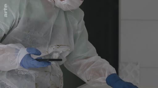 Un enquêteur de police scientifique, vêtu d'une combinaison de protection blanche et de gants en latex bleus, examine un téléphone portable sur une scène d'investigation.