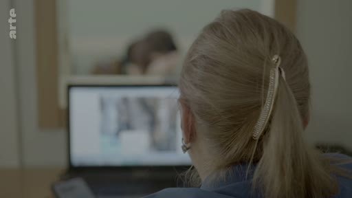 Une femme de dos, portant une pince à cheveux, consulte un écran d'ordinateur dans ce qui semble être un bureau ou un environnement administratif, dans le cadre d'une enquête sur l'identification de victimes.