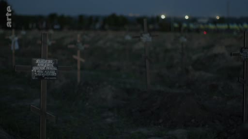 Vue d'ensemble d'un cimetière avec des croix en bois sur des tombes, filmée au crépuscule. Ce lieu de sépulture évoque le contexte de conflits ou de commémorations funéraires.