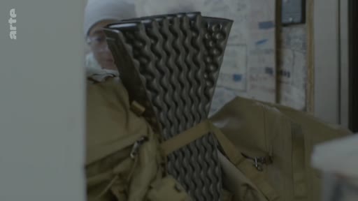Un soldat en tenue de camouflage prépare son équipement, incluant un matelas de sol en mousse, avant un départ en mission ou un retour au foyer.