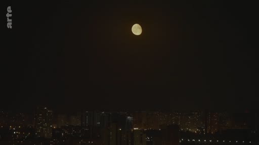 Vue nocturne d'une ville avec une pleine lune visible dans le ciel sombre au-dessus des immeubles éclairés.
