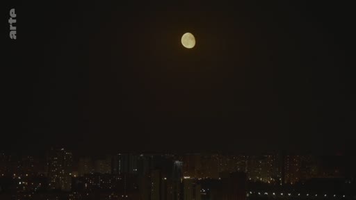 Vue nocturne d'une ville avec une pleine lune visible dans le ciel sombre au-dessus des immeubles éclairés.