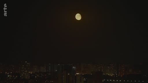 Vue nocturne d'une ville avec une pleine lune visible dans le ciel sombre au-dessus des immeubles éclairés.