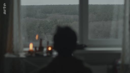 Une silhouette humaine est vue de dos, assise dans une pièce sombre devant une fenêtre donnant sur un paysage boisé, avec des bougies allumées sur le rebord. Cette image évoque une atmosphère de recueillement, de deuil ou de solitude.