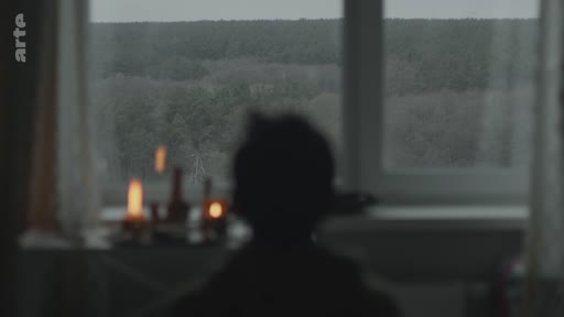 Une silhouette d'enfant est assise devant une fenêtre, observant un paysage forestier sombre à la lueur de bougies posées sur le rebord. Cette scène évoque une atmosphère de recueillement, de deuil ou de solitude.