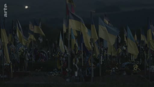 Vue d'ensemble d'un cimetière militaire en Ukraine, où de nombreux drapeaux ukrainiens flottent sur les tombes des soldats tombés au combat, sous une lune visible dans le ciel nocturne.