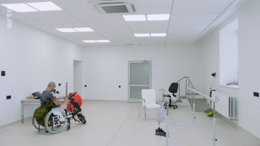 Un patient en fauteuil roulant équipé d'un dispositif de mobilité se trouve dans une salle de rééducation fonctionnelle moderne. La pièce est équipée de matériel médical, incluant des chaises et des barres parallèles pour la physiothérapie.