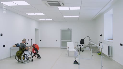 Un patient en fauteuil roulant équipé d'un dispositif de mobilité est en séance de rééducation dans une salle spécialisée avec des barres parallèles et du matériel médical.