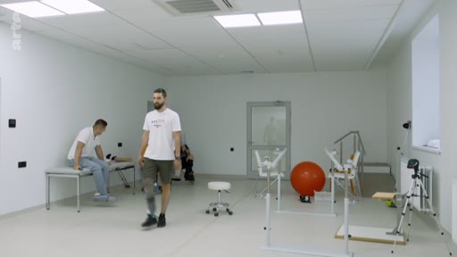 Un homme amputé équipé d'une prothèse de jambe effectue des exercices de marche dans une salle de kinésithérapie et de rééducation fonctionnelle. D'autres patients sont présents en arrière-plan dans ce centre de soins spécialisé.