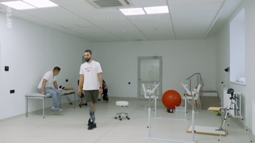 Un homme amputé d'une jambe marche dans une salle de rééducation équipée de matériel médical, accompagné par un kinésithérapeute. Cette scène illustre le processus de réadaptation physique et le travail de rééducation fonctionnelle dans un centre spécialisé.