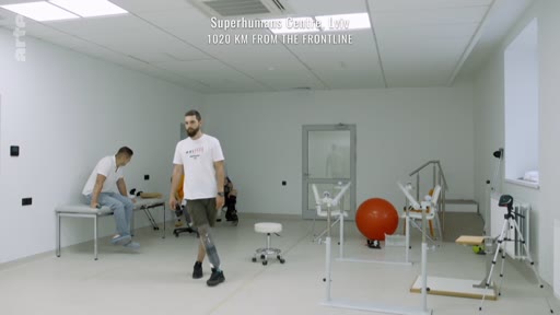 Un patient amputé équipé d'une prothèse de jambe effectue des exercices de marche dans une salle de rééducation du centre Superhumans, situé à 1020 km du front en Ukraine. Un autre homme est assis sur un banc dans cette salle équipée de matériel médical et de rééducation.