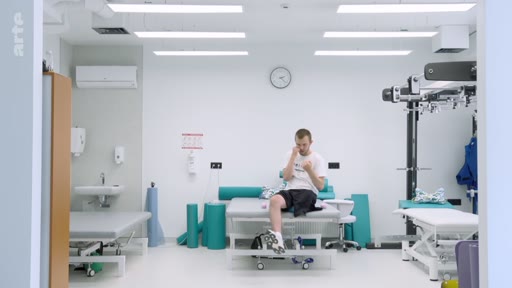 Un homme est assis sur une table d'examen dans une salle de kinésithérapie ou de rééducation fonctionnelle, équipée de matériel médical et de tapis de sport.