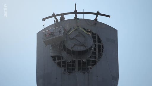 Une nacelle suspendue est utilisée pour démonter l'emblème soviétique, composé de la faucille et du marteau, situé au sommet d'un monument. Cette opération de décommunisation illustre le retrait des symboles de l'ère soviétique dans l'espace public.