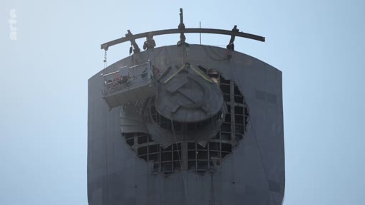 Une nacelle de chantier est utilisée pour démonter l'emblème soviétique, représentant la faucille et le marteau, au sommet d'un monument imposant. Cette opération symbolise la décommunisation et la suppression des vestiges de l'ère soviétique.