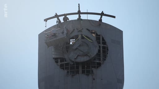 Une nacelle de chantier est utilisée pour démonter l'emblème soviétique de la faucille et du marteau situé sur la façade d'un monument imposant. Cette opération symbolique marque la décommunisation et le retrait des vestiges de l'ère soviétique.